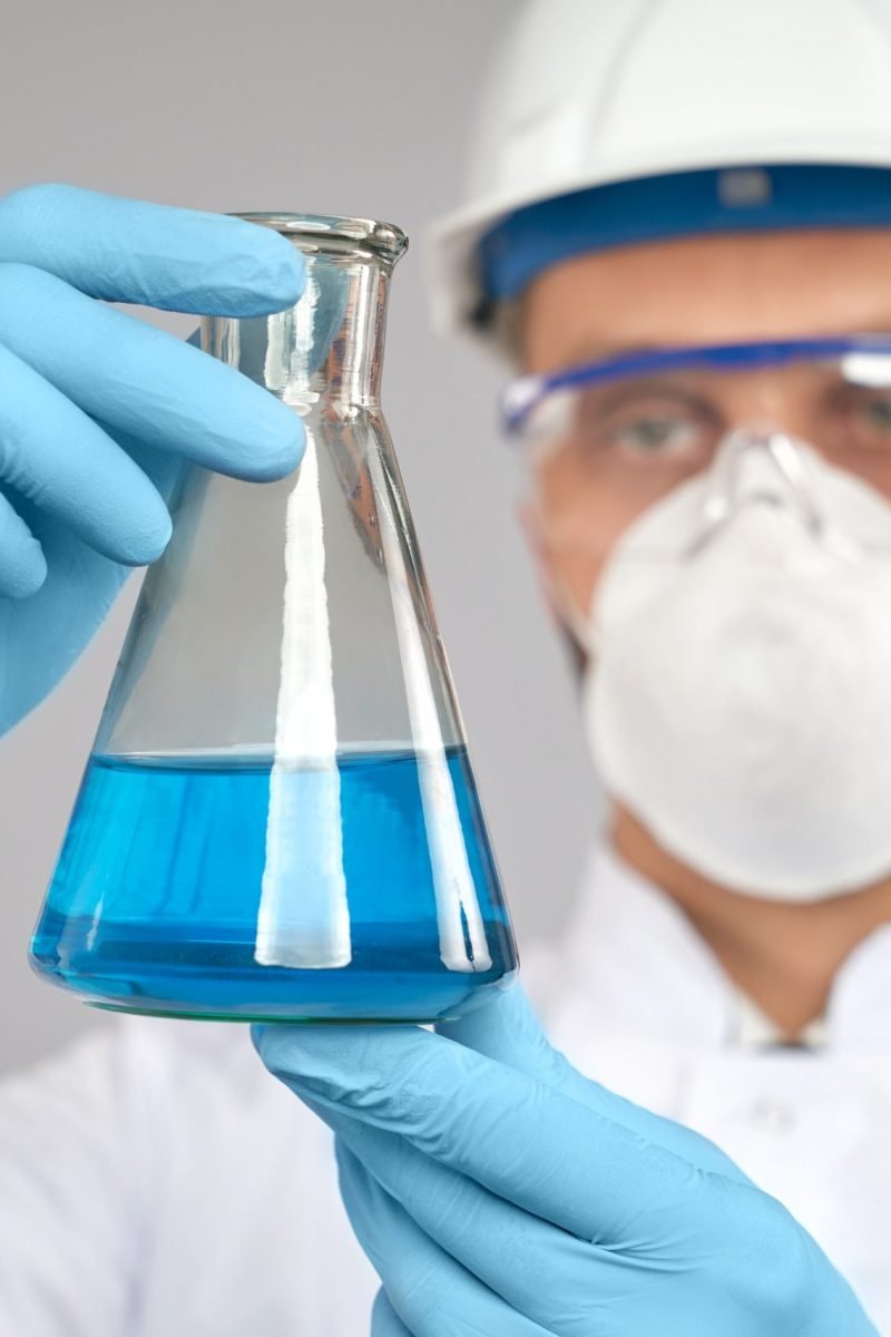 Close up of young chemist in glasses, helmet and mask doing experiment in laboratory, isolated on grey. Selective focus of flask with blue liquid in hand of scientist in gloves and white lab coat.